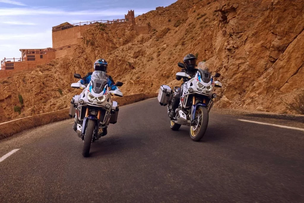 Dois pilotos testando a CRF1100L Africa Twin 2024 em uma estrada estreita e asfaltada em uma montanha do deserto.
